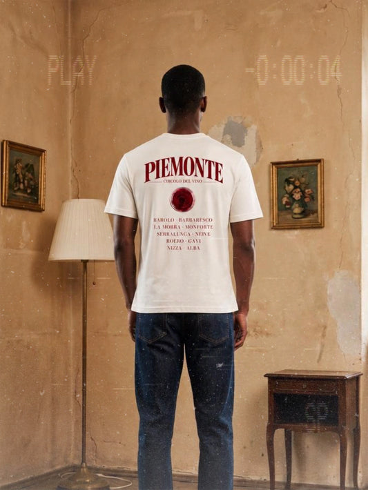 Man wearing a BACCONE Piemonte Circolo del Vino premium T-shirt in vintage white with burgundy red wine-inspired back print, standing in a rustic Italian-style interior.