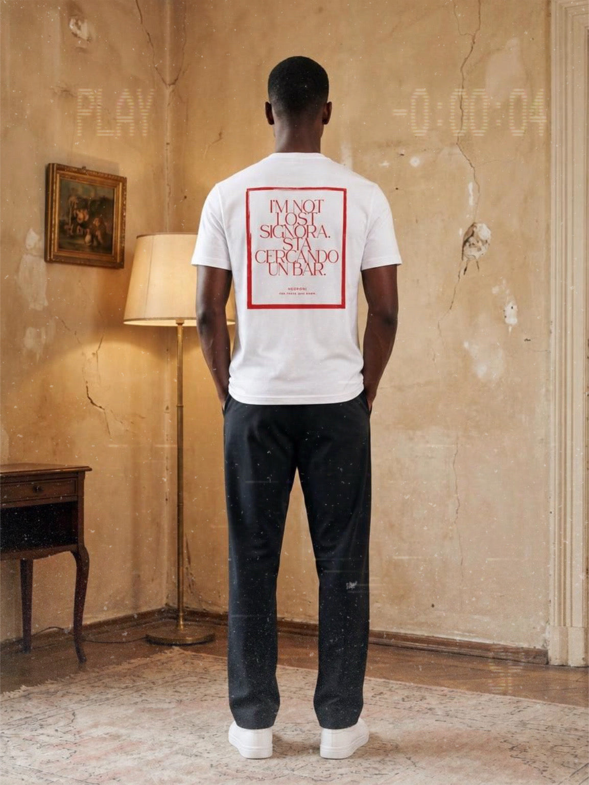 Man wearing a white BACCONE Cercando Bar organic cotton T-shirt with red "I'm Not Lost, Signora. Sto Cercando Un Bar." back print, standing in a vintage Italian-inspired interior.