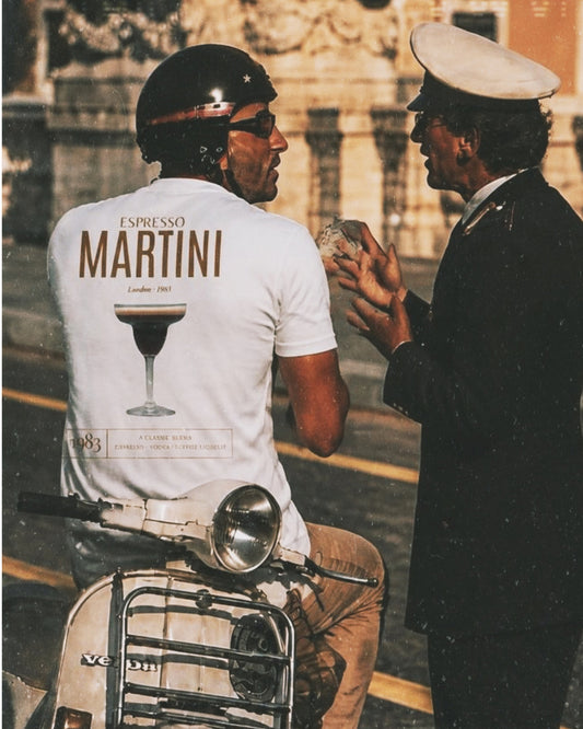Man wearing a white BACCONE Espresso Martini premium T-shirt with vintage cocktail back print, sitting on a Vespa and talking to a gentleman in a nostalgic Italian street setting.