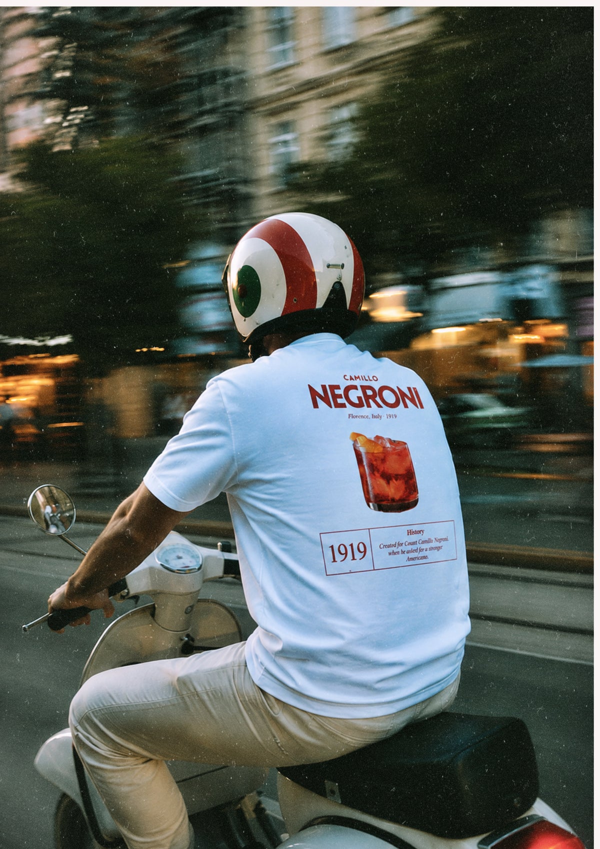 BACCONE Negroni Il Classico premium white T-shirt with red Negroni cocktail back print, worn by a man riding a Vespa in an Italian street-style setting.