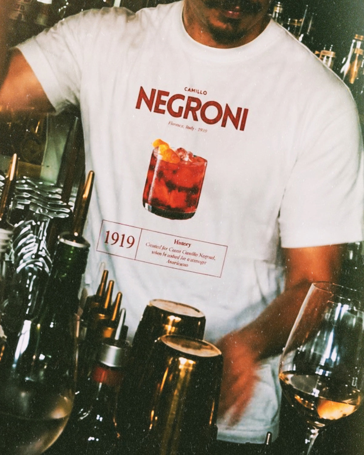 BACCONE Negroni Il Classico T-Shirt in Weiss mit rotem Cocktail-Frontprint, getragen von einem Barkeeper in einer italienisch inspirierten Bar.